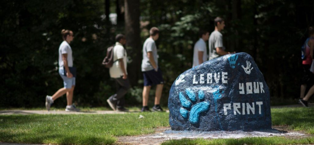 A painted rock with "Leave Your Print" and a paw print is in the foreground, while a group of people walk along a wooded path in the background.