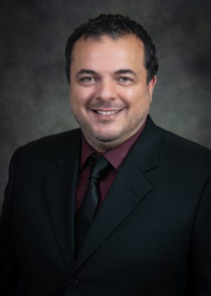 Man in a dark suit, maroon shirt, and black tie, smiling, with a dark textured background.