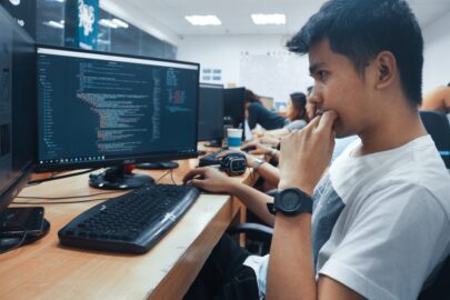 A person sits at a desk in an office, focused on a computer monitor displaying lines of code, embodying the spirit of computer science. With a keyboard and mouse in front, several other individuals work diligently in the background.