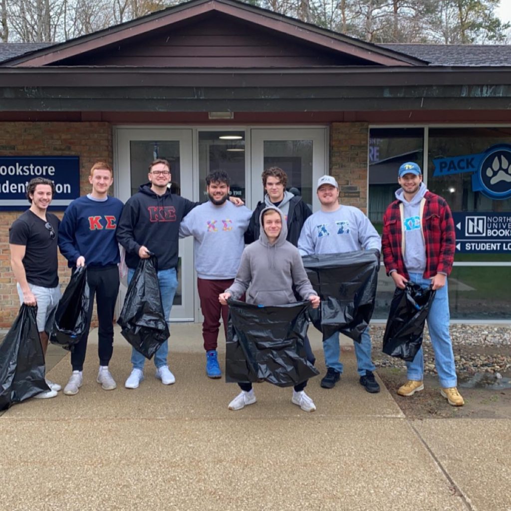 A group of seven young men from Kappa Sigma, wearing casual clothes and holding black trash bags, stands in front of a bookstore and student union building.