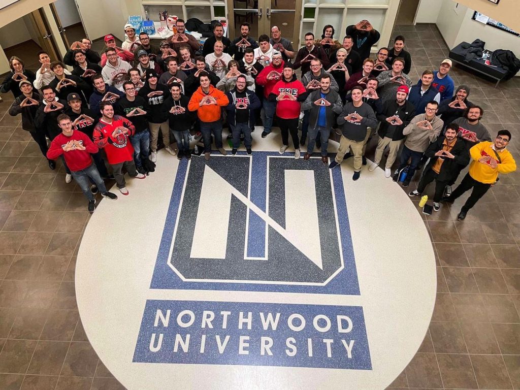 A large group of people stand in a circle around a Northwood University logo on the floor, many making triangle hand signs associated with Tau Kappa Epsilon, in a lobby area.
