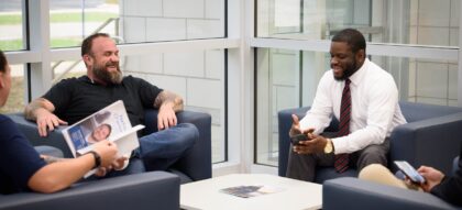 Four men are seated in a modern office lounge area, two engaged in conversation, with one reading a brochure about the Master of Business Administration program and another looking at his phone.