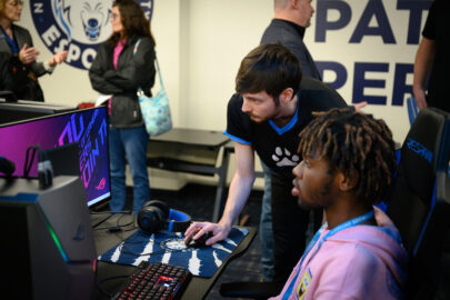 One student in a hoodie sitting in a gaming chair while another Esports student controls the mouse of the computer they are both looking at.