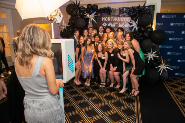 A group of people poses for a photo at the Auto Show Gala with a Northwood-themed backdrop and decorations, while another person stands at a photo booth setup.
