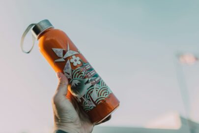 Hand holding an orange water bottle with a silver cap and patterned design against a light sky background.