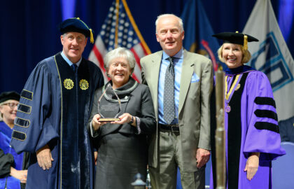 Four people, three in academic regalia, and one holding a trophy, stand together smiling in front of flags on a stage.
