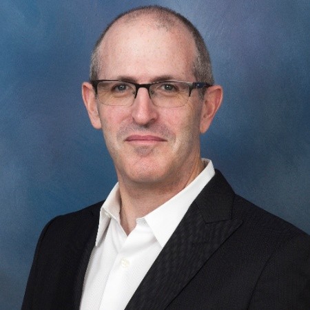 A man with glasses wearing a white shirt and a black blazer poses against a blue background.