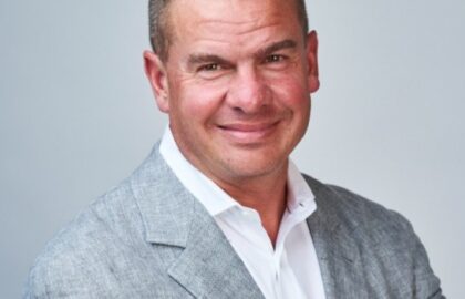 A man in a light gray suit jacket and white shirt smiles with arms crossed, exuding leadership insights against a plain background.