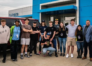 A group of people stands together outside a blue and white building, posing for a group photo on a cloudy day.