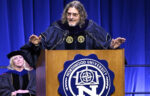 A man in academic regalia speaks at a podium with the Northwood University emblem, demonstrating leadership to the Class of 2025, while a woman in similar attire sits behind him on stage.