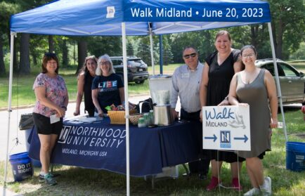 Seven people stand under a Northwood University tent with a "Walk Midland" sign and table of refreshments, outdoors on June 20, 2023.