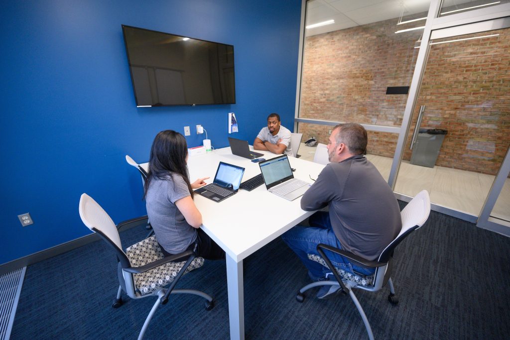 Three people sit around a table with laptops in a modern conference room, discussing their MS in Business Analytics projects. The room features a large screen on the wall and a glass partition, providing an ideal setting for collaboration and data-driven decision-making.