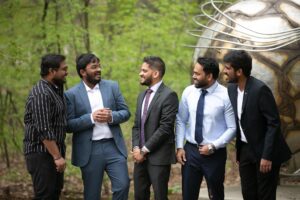 Five men in formal and casual attire stand together outdoors, smiling and talking near a large metallic sculpture.